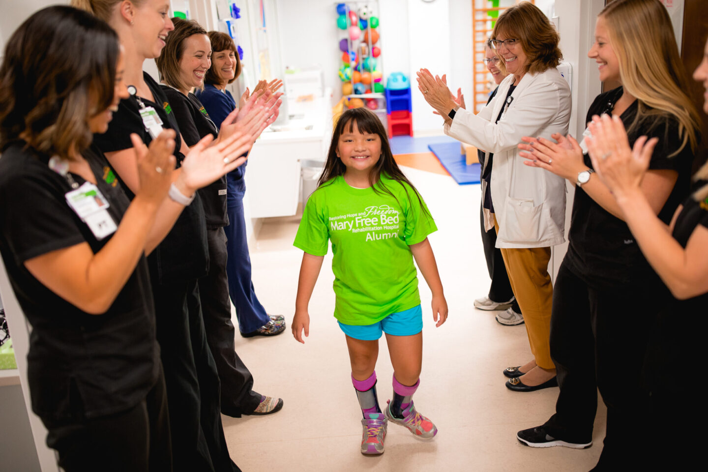 A young girl with leg braces walks confidently through a hallway as staff applaud her progress