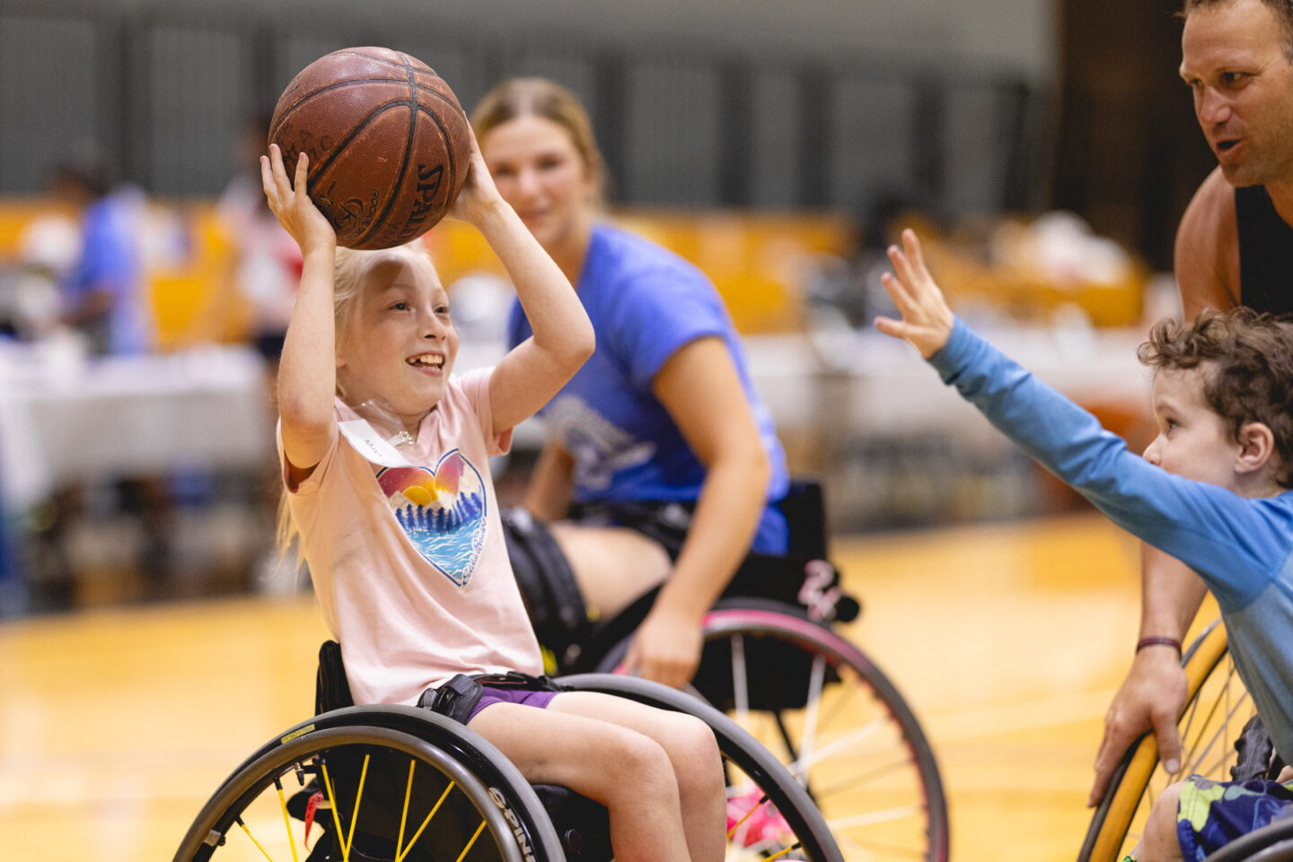 A young girl in a wheelchair smiling and holding a basketball above her head during the Junior Wheelchair Sports Camp.
