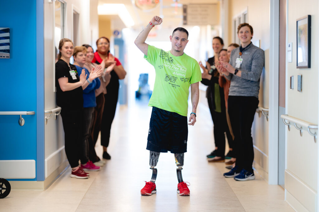 A man with double leg prosthetics triumphantly raises his fist while walking down a hallway lined with applauding staff at Mary Free Bed.