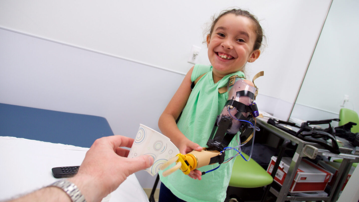 Child smiling and using her prosthetic arm to grab a paper cup from a clinician.