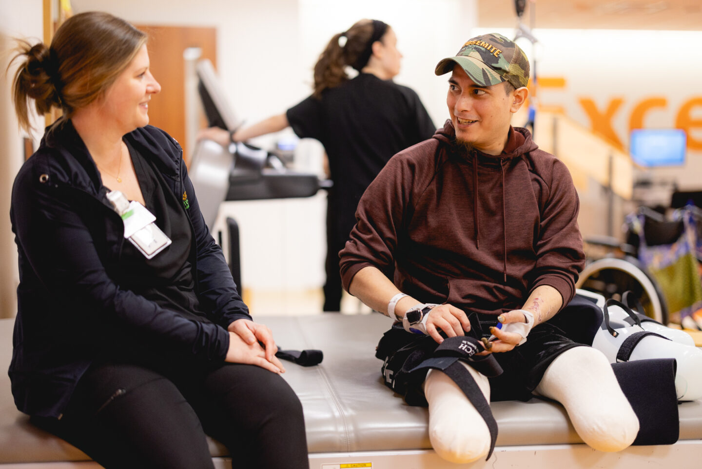 A patient with bilateral leg amputations sits on a treatment table, engaging in a conversation with a physical therapist.