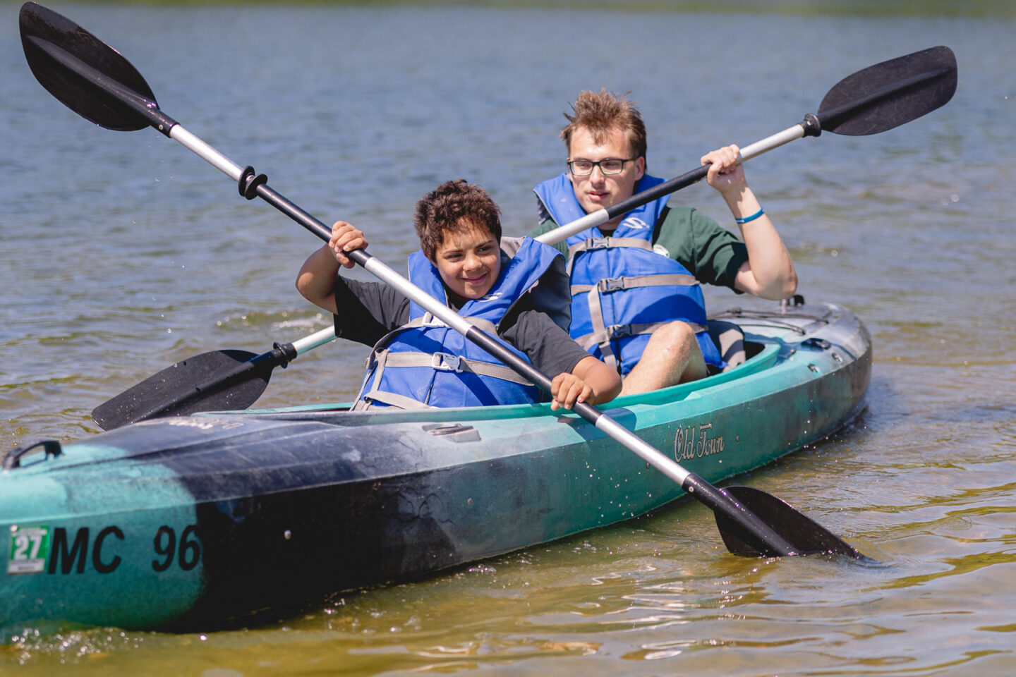 Two individuals in blue life jackets paddling a kayak on a lake, participating in an adaptive sports clinic at Millennium Park.