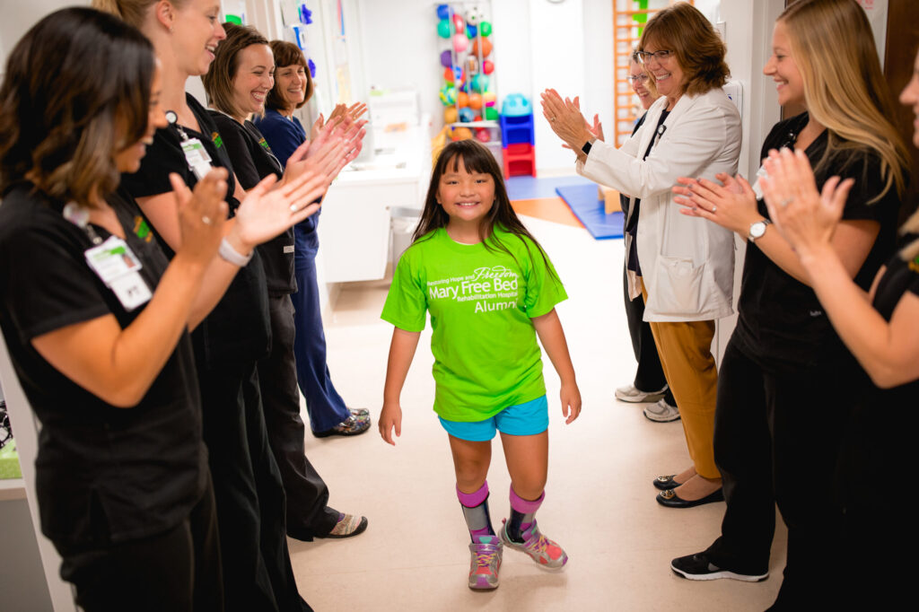 A young girl with leg braces walks confidently through a hallway as staff applaud her progress.