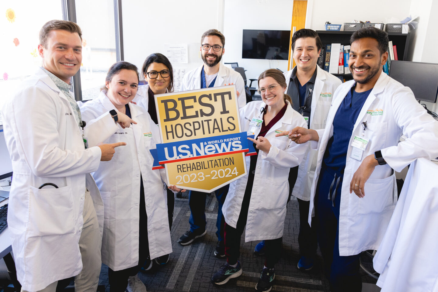 A group of seven residents in white lab coats are standing together in a room, smiling and holding a large sign that reads "BEST HOSPITALS, U.S. News & World Report, Rehabilitation 2023-2024" The individuals are standing close together, some pointing at the sign, celebrating their achievement