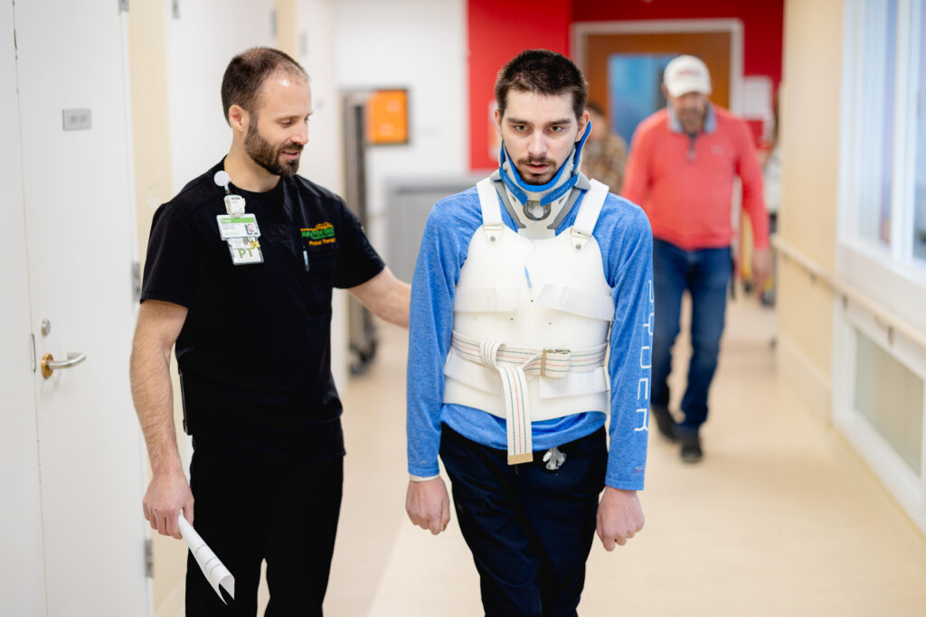 Male patient wearing a spinal brace walking with guidance from a physical therapist.