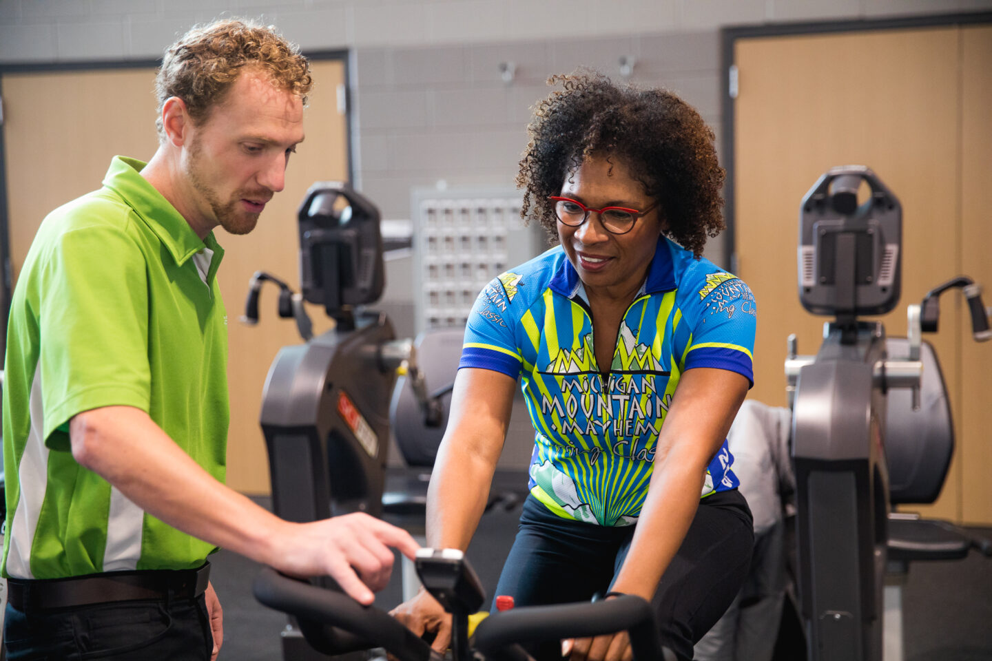 A physical therapist assists a patient on a stationary bike at the Mary Free Bed YMCA. The patient, wearing a Michigan Mountain Mayhem cycling jersey, is engaging in low-impact cardiovascular exercise as part of their sports rehabilitation program.