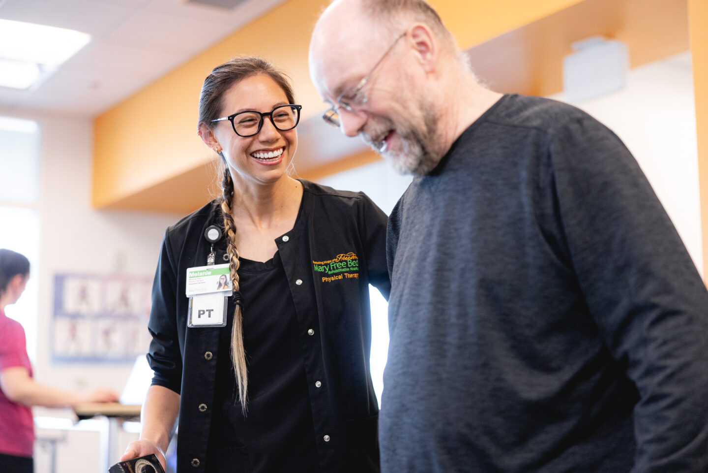 A physical therapist smiles and supports an older adult patient during a rehabilitation session at the SpartanNash YMCA. The patient is walking with the therapist’s guidance, focusing on mobility and balance as part of his recovery process in a sports rehabilitation program.