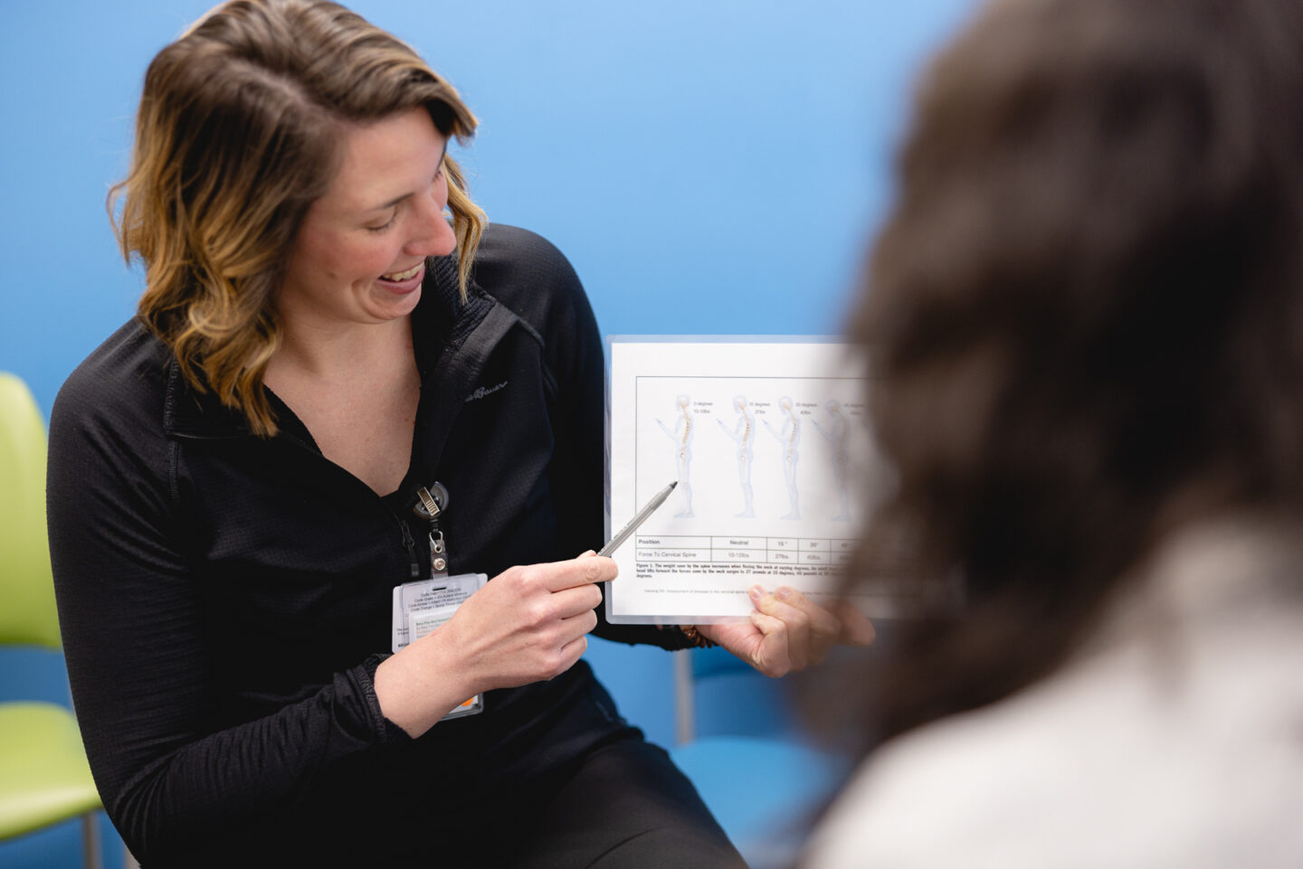 A physical therapist reviews a spine alignment chart with a patient during a sports rehabilitation consultation at the SpartanNash YMCA. The therapist points to different spinal positions, discussing posture and injury recovery strategies.