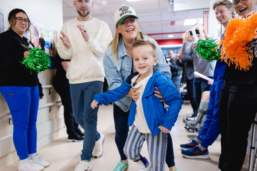 Young child celebrating with healthcare staff in a graduation tunnel decorated with green and orange pom-poms, marking a significant milestone in pediatric rehabilitation