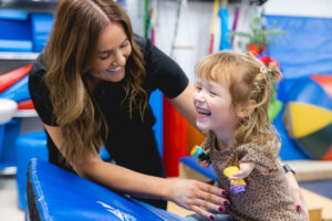 Pediatric occupational therapist engaging with a young child in a bright therapy gym, guiding fine motor and coordination activities using colorful equipment and therapeutic tools