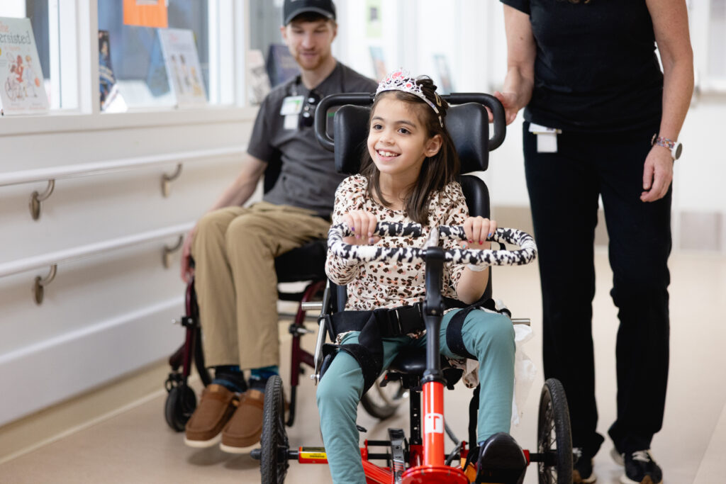 Young child using a red adaptive tricycle with support from recreational therapy staff in a clinical rehabilitation setting, promoting mobility and independence
